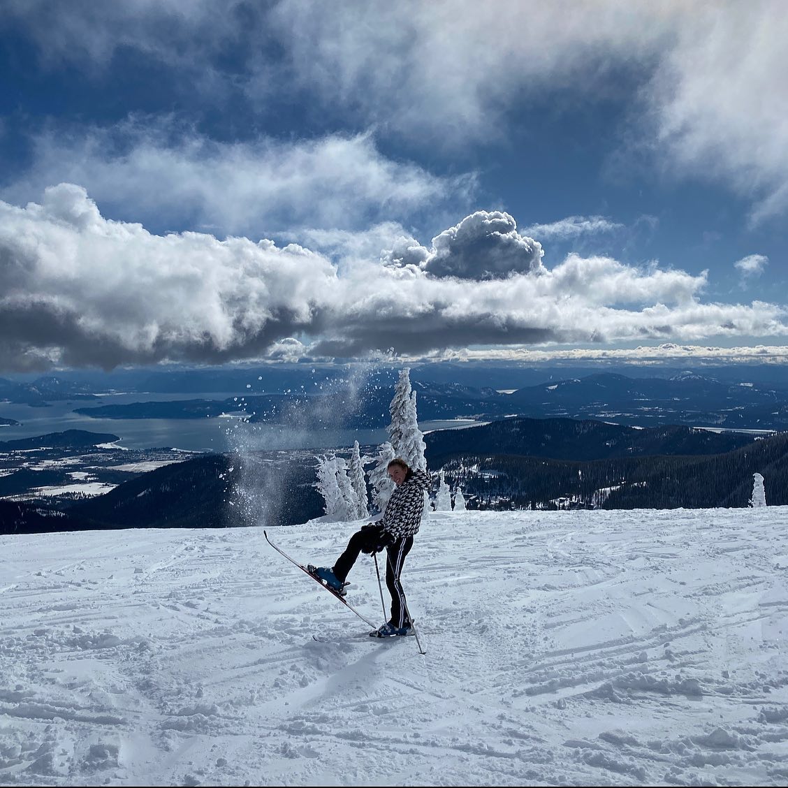 a person is skiing down a snowy mountain slope. the person is wearing a black and white jacket and is kicking one ski up in the air. there is snow spraying up from under their skis. the background shows a stunning view of a valley with a lake in the distance, surrounded by snowcapped mountains under a partially cloudy blue sky. the scene is very picturesque and shows a beautiful winter landscape.