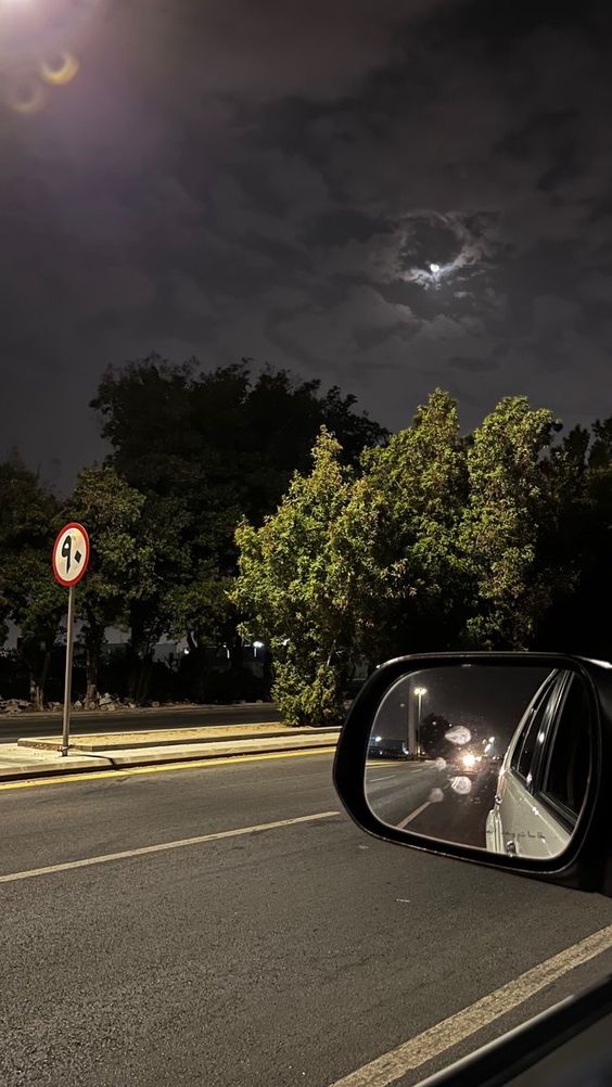 the image shows a view from the driver's seat of a car at night. the side view mirror reflects the road behind, illuminated by streetlights and the moon partially hidden behind clouds.  trees line the side of the road, and a street sign is visible in the distance. the overall atmosphere is dark and moody, with a sense of movement implied by the car's perspective.