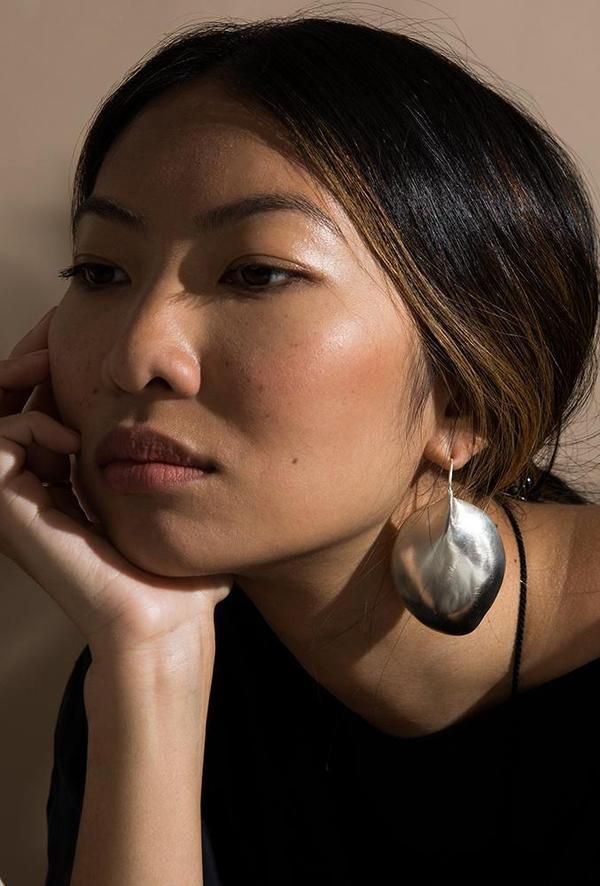 a closeup portrait of a woman with dark hair and a serious expression, wearing a large silver earring. her hand is on her face, and she's wearing a black top.