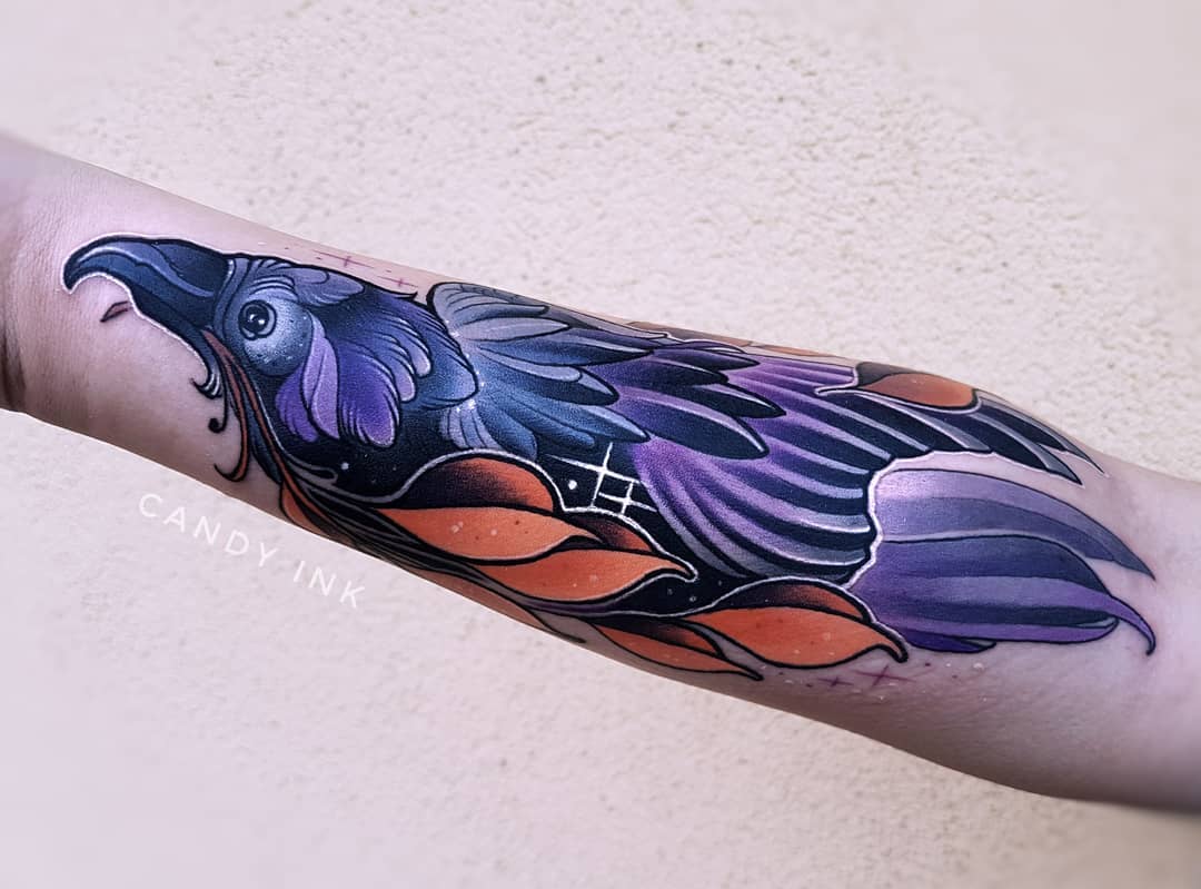 a closeup photograph showcases a freshly done raven tattoo on someone's inner forearm. the tattoo depicts a raven in flight, rendered in a neotraditional style. the bird's plumage displays a captivating blend of deep purples and blues, accented with vibrant orange feathers that resemble flames. the intricate details of the feathers and the bird's intense gaze create a striking visual impact.
