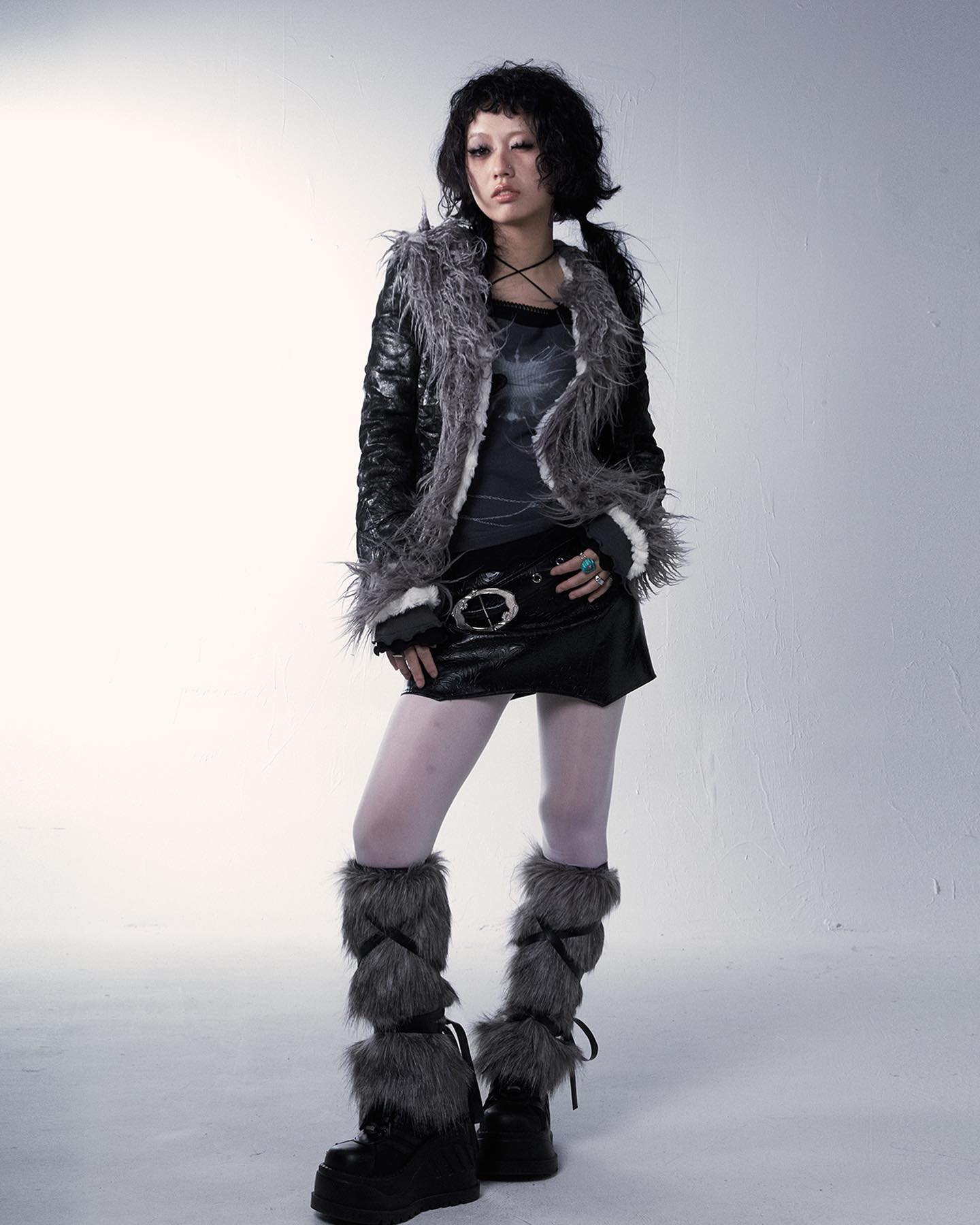 a fullbody studio shot of a young asian woman modeling a stylish outfit. she's wearing a gray fauxfur trimmed black leather jacket, a black miniskirt, white tights, and gray fauxfur leg warmers with black boots. her dark hair is styled short. the overall style is edgy, blending punk and alternative elements. the background is a simple, neutral white. the model is posing slightly angled, with a hint of attitude in her expression.