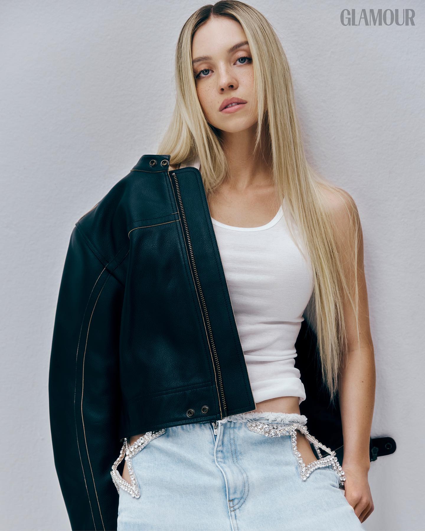a young blonde woman is featured in a glamour magazine photoshoot.  she's wearing a black leather jacket over a white tank top and light wash denim shorts adorned with rhinestone embellishments forming a star pattern along the sides. the photo is a closeup studio shot, showcasing her style and the details of her outfit. the background is a plain, light gray.