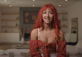closeup shot of a young, attractive redhead woman with long, curly hair. she's smiling and looking directly at the camera. she's wearing a red polka dot offtheshoulder crop top and a necklace. the background is blurred but shows a living room setting. the overall mood is relaxed and casual.