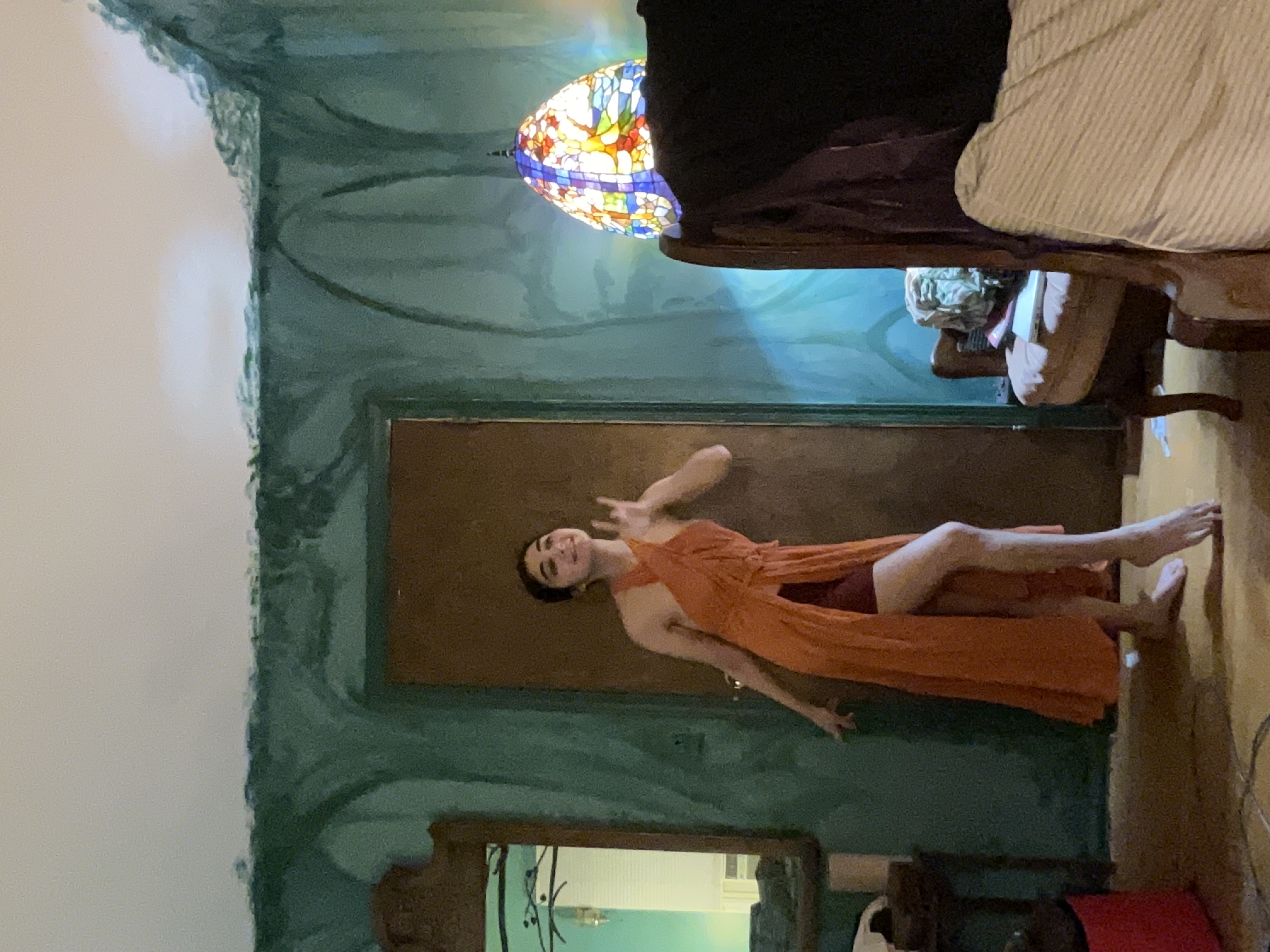 a young woman in an orange wrap dress with a high slit is standing in a room with green walls. she is posing with one hand on her hip. the room features an antique stainedglass lamp, an old chair, and a mirror. the overall style is vintage or retro.