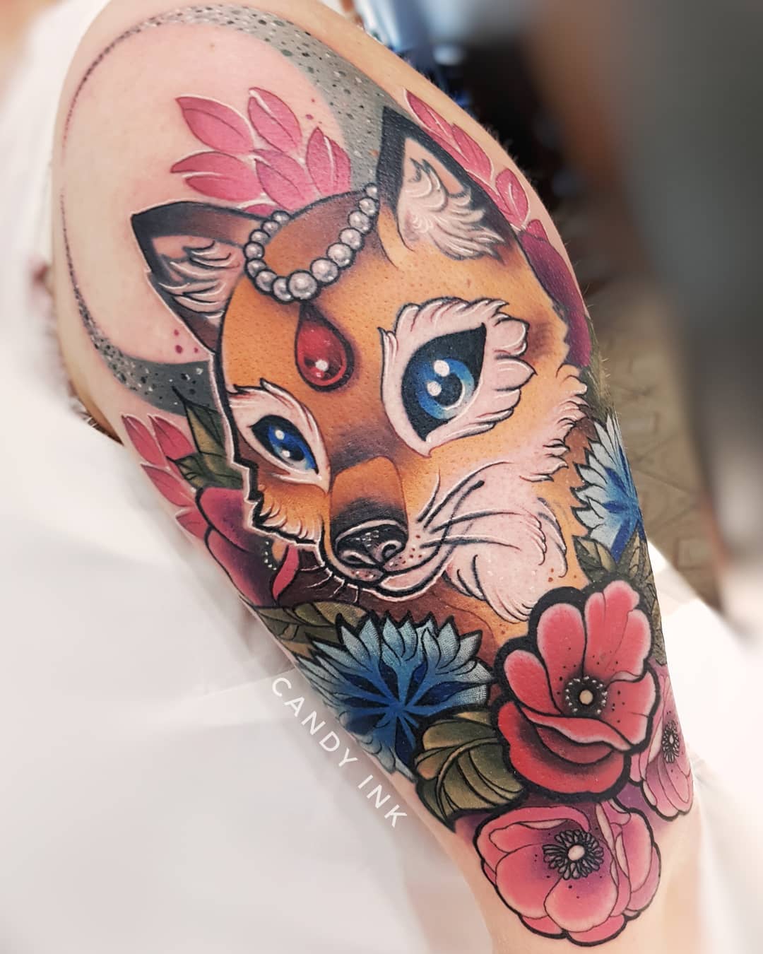 a person's upper arm with a colorful tattoo of a fox surrounded by flowers. the fox has blue eyes, a red jewel on its forehead and a string of pearls in its hair.