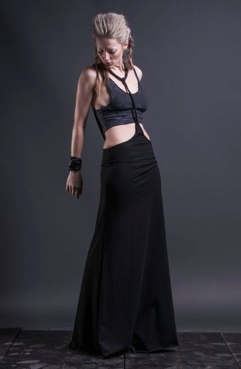 a fulllength studio shot of a woman wearing a dark outfit against a dark gray background. she has a modern, edgy style. the woman is looking down with her head slightly tilted. her blonde hair is partially braided and styled with some volume at the front. she is wearing a dark, sleeveless crop top paired with a highwaisted black maxi skirt. the skirt extends to the floor, hiding her shoes. the outfit features suspenderlike straps that connect the skirt to the top. she also wears a thick bracelet on her left wrist.