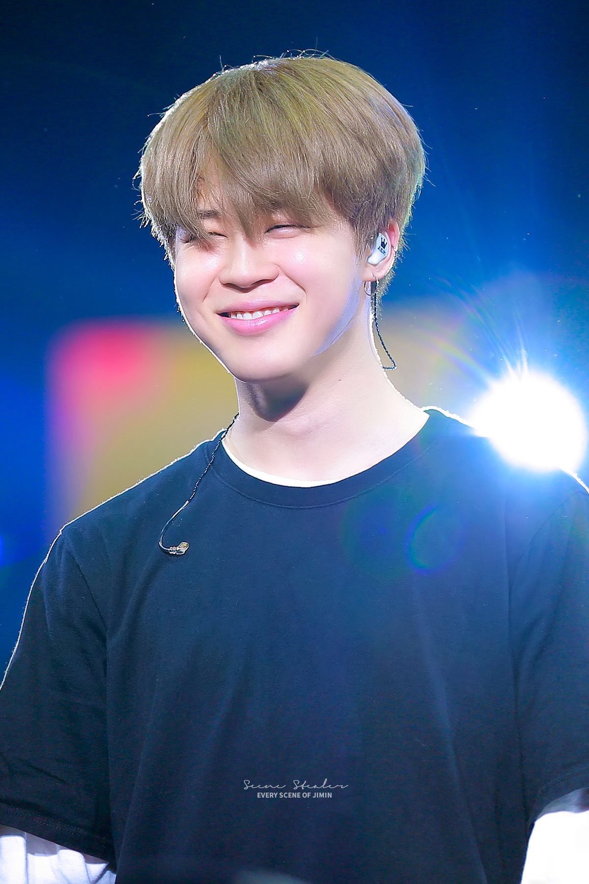 a photo of park jimin from bts smiling onstage wearing a black shirt with a white undershirt, blond hair, and inear monitors.