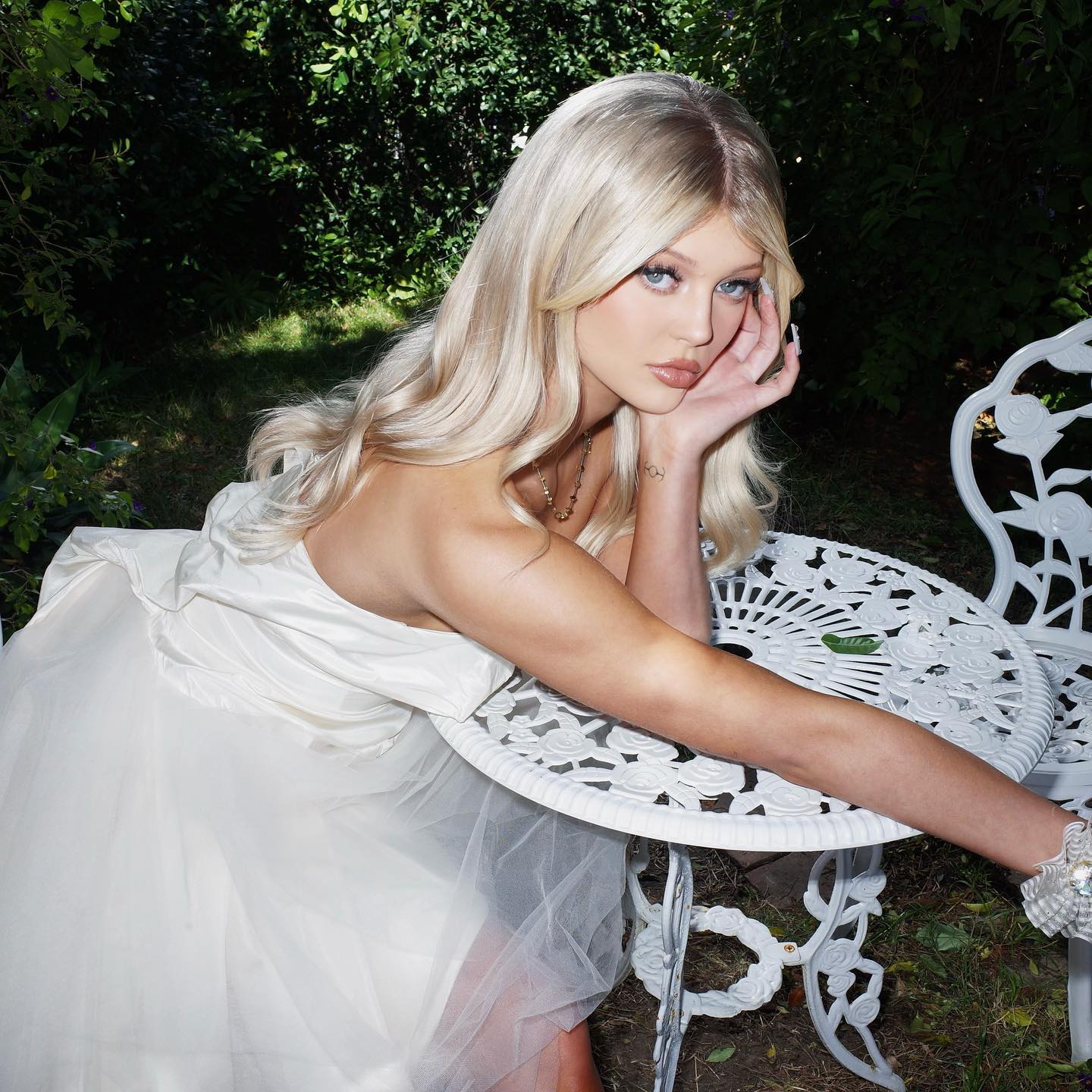 a woman with blonde hair is wearing a white dress and jewelry while leaning on a white table outdoors in a garden. she is posing for a portrait.