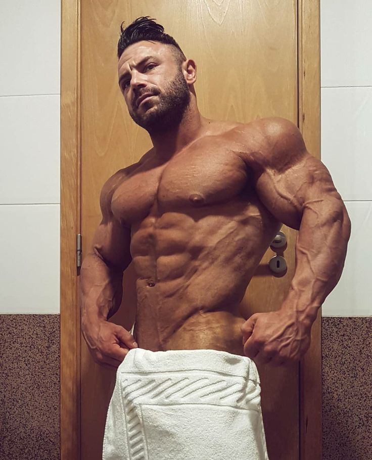 the image shows a muscular, tanned man with a welldefined physique. he appears to be a bodybuilder, showcasing highly developed pectoral muscles, abdominal muscles (sixpack), and prominent veins. he's posing in a bathroom, partially wrapped in a white towel, against a wooden door. the lighting is slightly dim, emphasizing the muscular definition.  his expression is serious and direct, looking toward the camera. the overall impression is one of strength, fitness, and possibly selfassuredness.