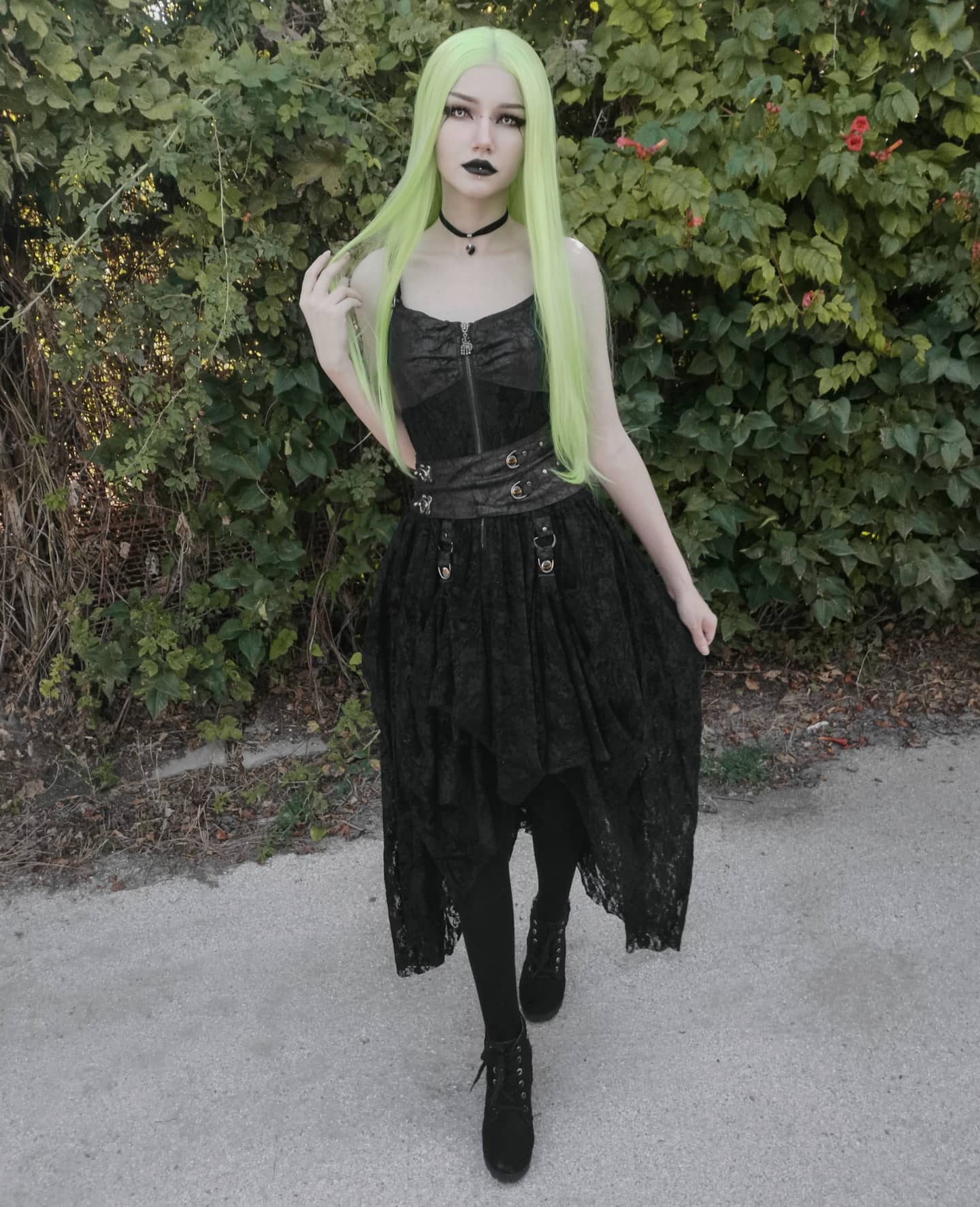 a young woman with vibrant pastel green hair is standing outdoors against a backdrop of lush green foliage. she is dressed in a stylish black lace dress with a corsetstyle top and a highlow skirt hemline.  the dress has decorative buckles and metallic accents, suggesting a steampunk or gothic influence.  she's wearing black boots and dark makeup, completing the dark and alternative fashion aesthetic. the overall impression is one of edgy and fashionable gothic style.