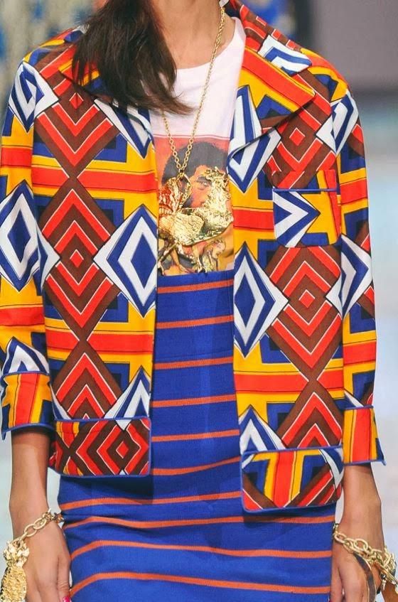 loseup view of a woman's vibrant outfit. she is wearing a shortsleeved jacket with a bold geometric pattern in shades of orange, red, yellow, blue, and white.  underneath, she is wearing a blue and orange striped skirt or dress.  the jacket has a relaxed, almost pajamalike style.  she is also wearing a gold necklace with a large pendant and gold bracelets. the overall style is bold and colorful, suggesting a runway or fashion show setting.  the image focuses on the texture and pattern of the clothing.