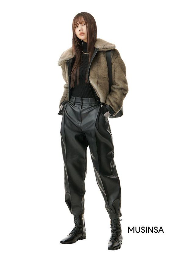 a young woman with long dark hair stands against a white background. she wears a brown fur jacket with a black turtleneck sweater underneath, black leather pants with pleats around the hips, and black combat boots. she gives a neutral expression and poses with her hands in her pockets. the image has a clean, studio aesthetic with bright artificial lighting.