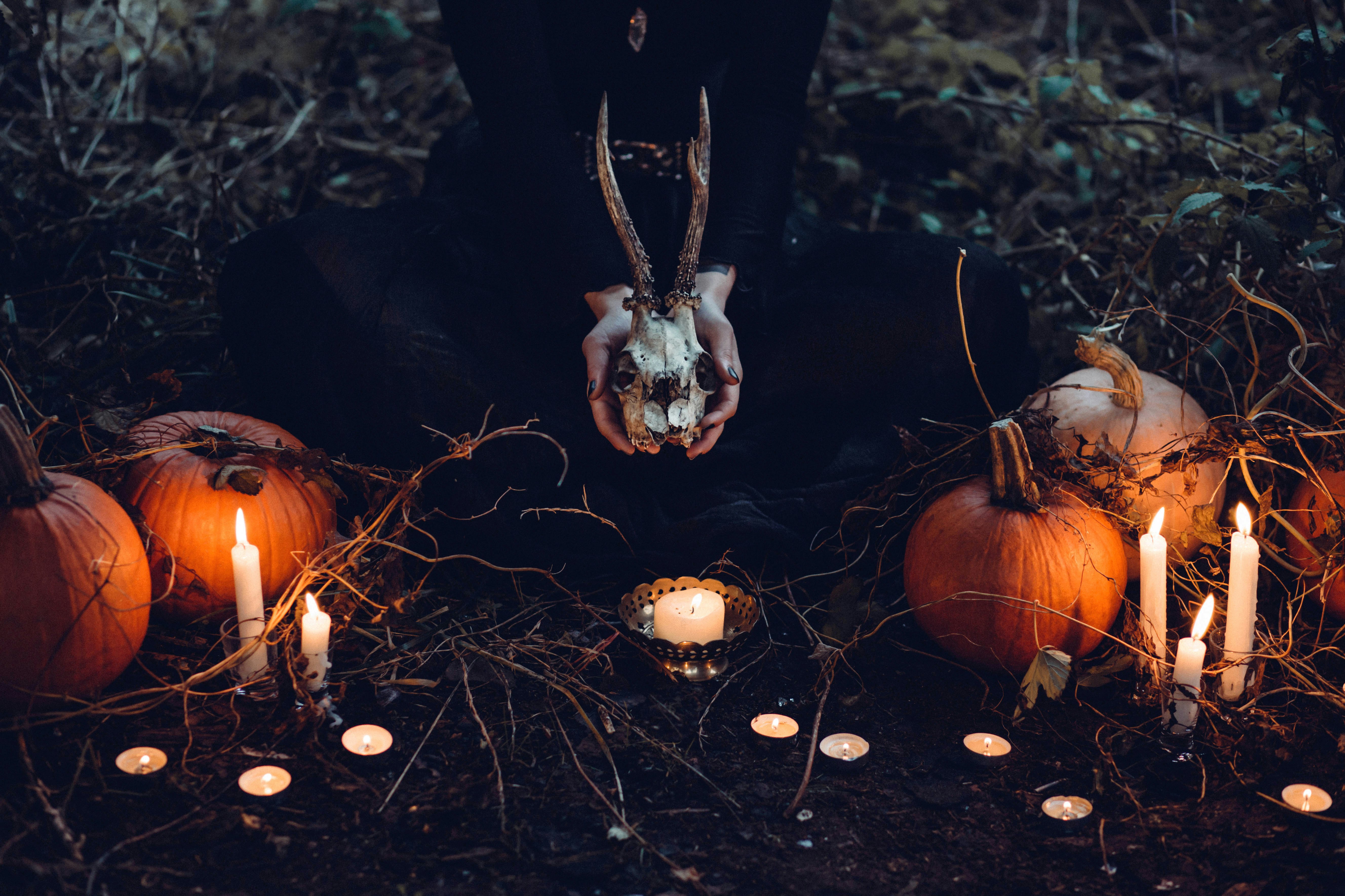 a person wearing a black dress sits in a forest surrounded by lit candles, pumpkins, and branches. in their hands, they hold a small animal skull. the scene evokes themes of halloween, witchcraft, and autumn.