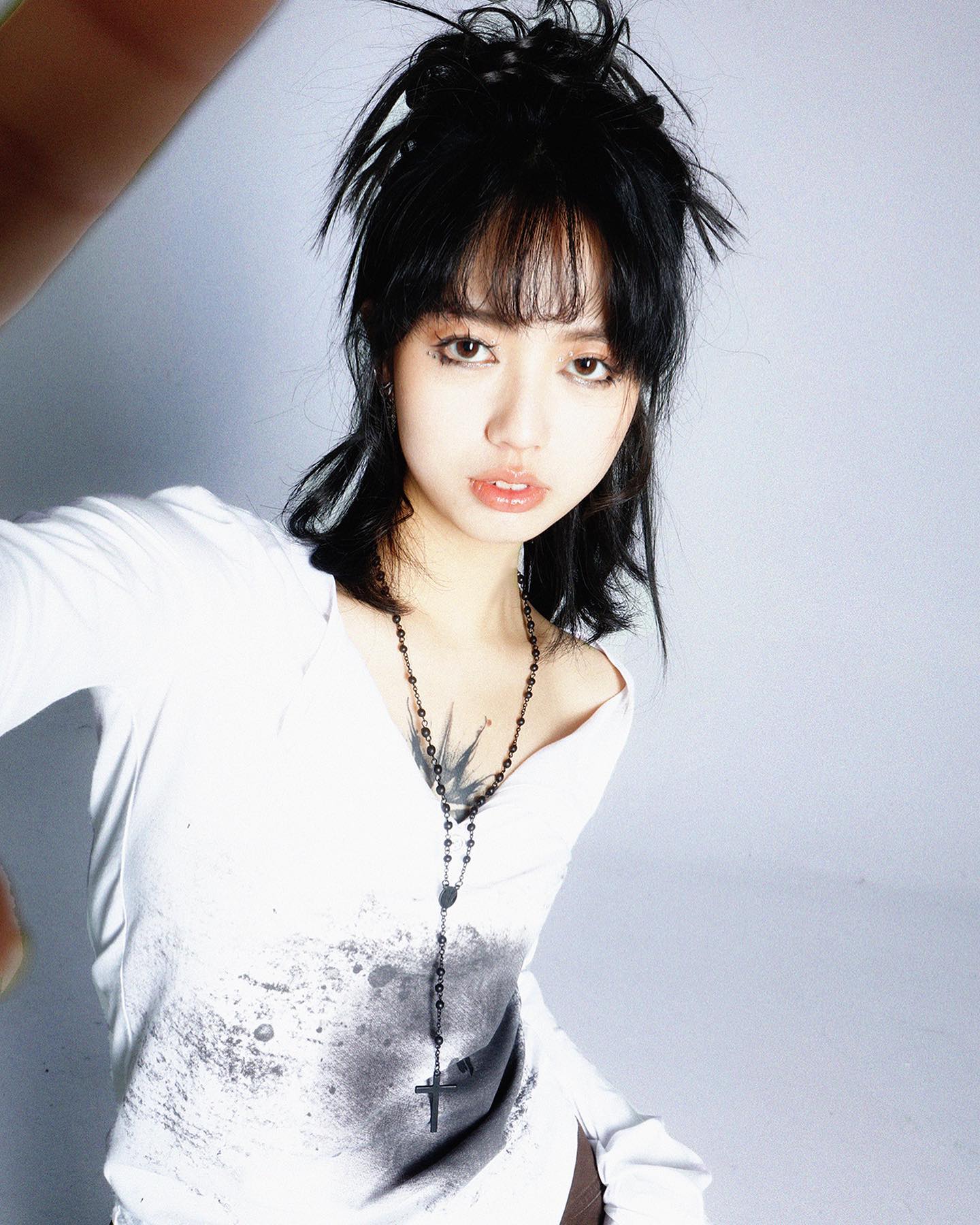 loseup portrait of a young asian woman with black hair styled with bangs and a messy top. she's wearing a white longsleeved shirt with a dark splatter pattern and a black rosary necklace with a cross. she has a tattoo visible on her chest. her expression is calm and she is looking directly at the camera. the background is a plain light grey. the overall style is a mix of casual and edgy.