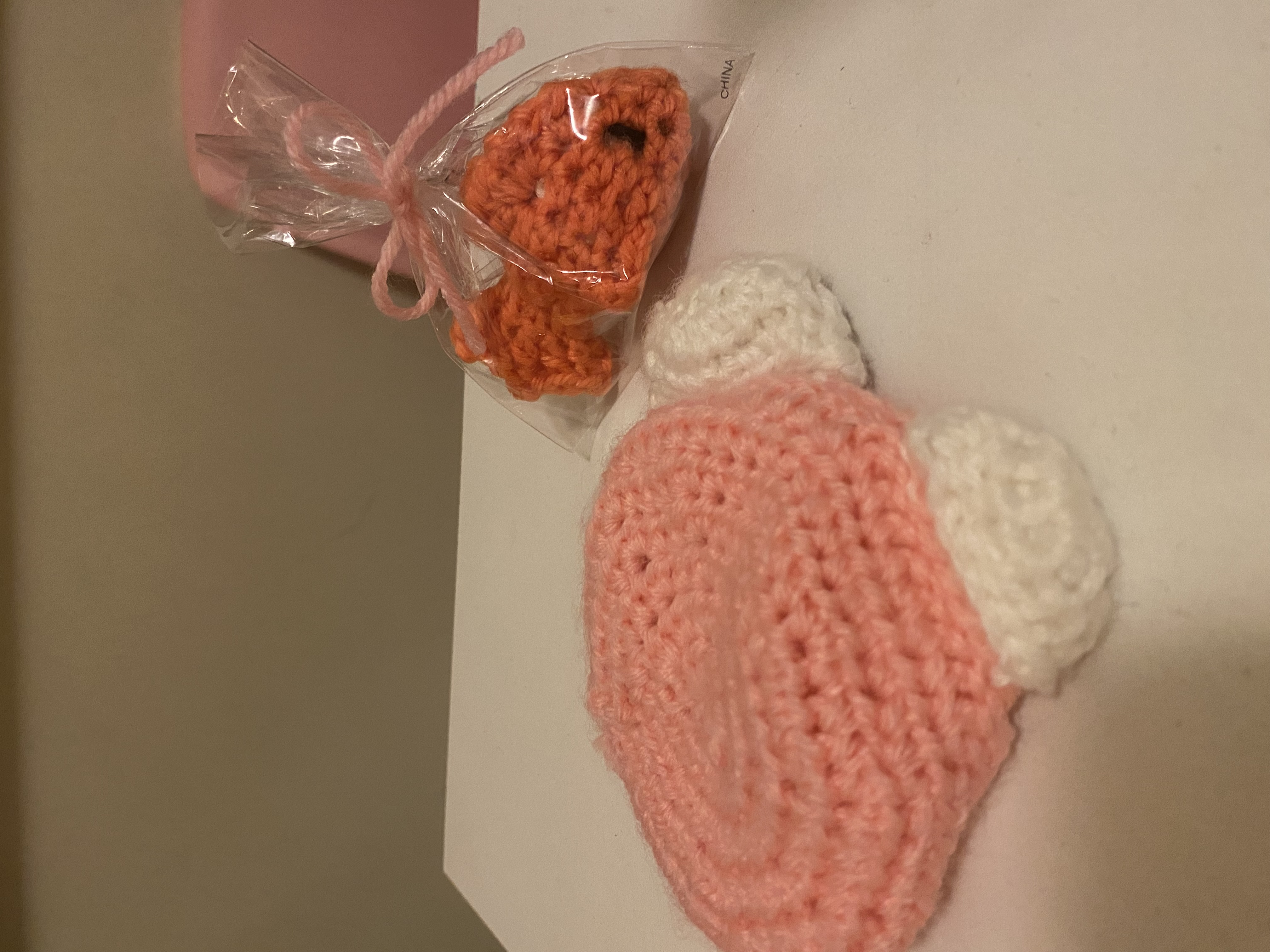 the image shows two small crocheted items on a white surface. one is a pink crocheted hat with white accents resembling a cartoonish character, possibly a baby's hat. the other is a small, orange crocheted fish, packaged in a clear plastic bag tied with a ribbon.  both items appear to be handmade and are likely toys or gifts. the background suggests a domestic setting.