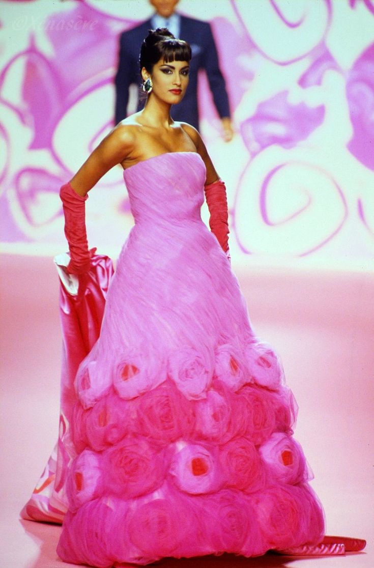 a woman in a pink, strapless gown is standing on a runway. the dress is layered with a gradient of pink roses near the bottom. she has pink, long gloves and wears earrings. a man is blurred in the background.