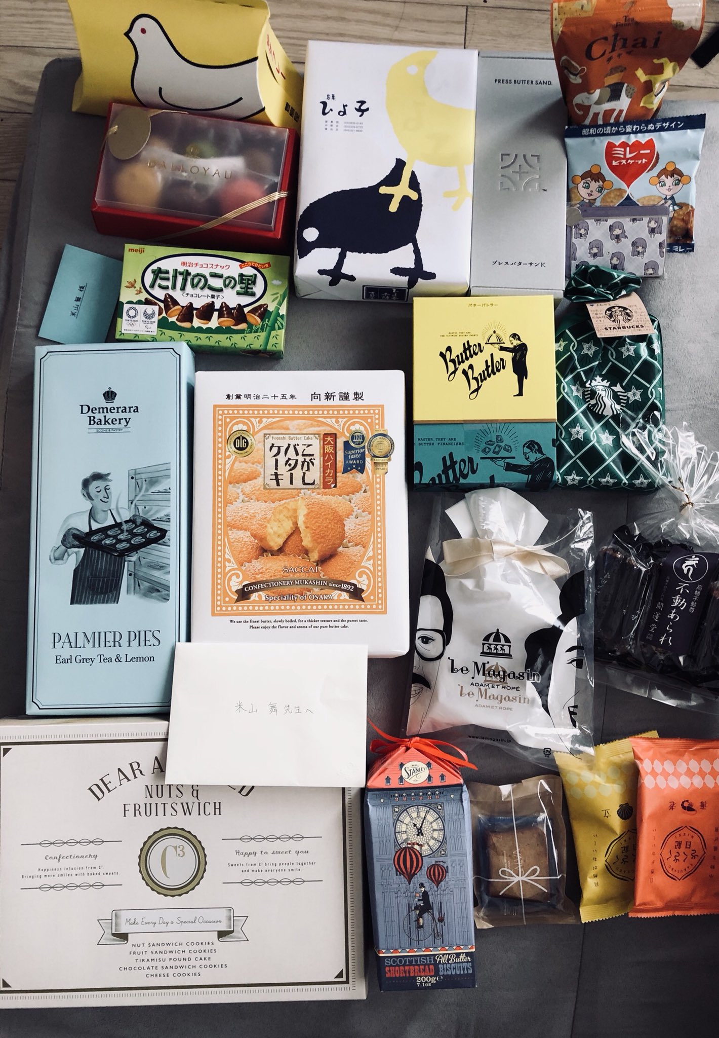 the image shows a collection of japanese snack boxes and souvenirs arranged on a beige surface, likely a bed or floor. there are various types of sweet treats, including cookies and chocolates. the packaging features whimsical designs with illustrations of birds, men, and hot air balloons. a handwritten card suggests the items might be gifts. the overall impression is one of delight and abundance, hinting at a thoughtful gesture or a successful shopping spree.
