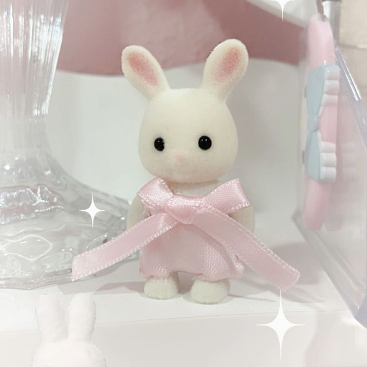 a white rabbit toy, possibly from the sylvanian families collection, is wearing a pink dress with a bow. the rabbit is standing on a white surface. to the left, there is a clear glass vase, and in the background, there are pastel pink and white elements. another miniature rabbit can be seen at the bottom left corner. the overall aesthetic is soft and pasteltoned.
