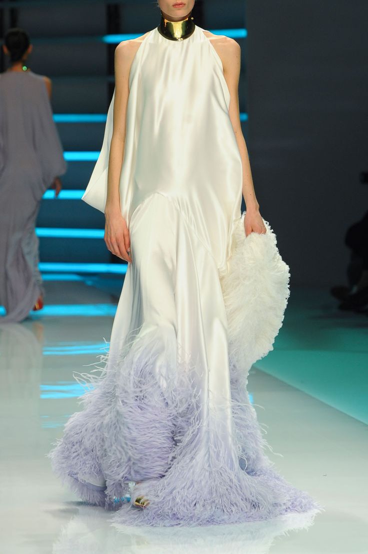 a fashion model walks the runway in a long, white satin dress with a feather trim. she is wearing a gold collar around her neck and her hair is styled in a sleek, straight style. the runway is set against a blue backdrop, with a white floor. the model is looking straight ahead, with a serious expression on her face. the dress is a beautiful example of high fashion, with its elegant silhouette and stunning feather trim. the model's accessories, including her gold collar, are also very stylish and add to the overall look. this is a beautiful and glamorous image of a fashion model on the runway.