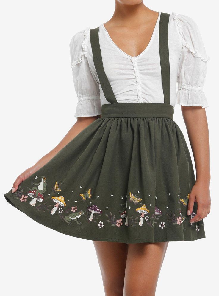 a woman models a green suspender skirt with embroidered mushrooms, frogs, and butterflies. the skirt falls above the knee and is paired with a white, peasantstyle top. the background is a plain white, suggesting a studio setting.