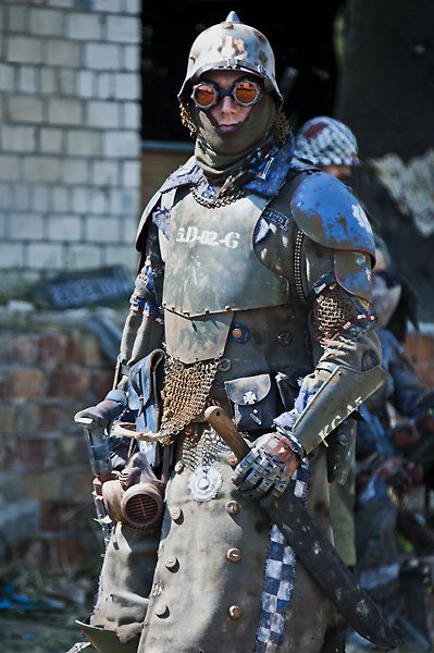 a person in elaborate postapocalyptic armor stands in front of a crumbling brick building. the armor is made of metal plates and chainmail, with a gas mask and goggles covering the face. the person is also armed with a large axe. the overall tone of the image is gritty and dystopian.