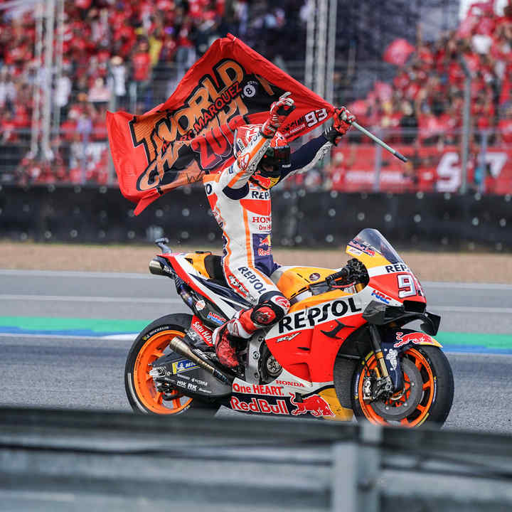 a photo of marc marquez celebrating his victory after a motorcycle race. he is riding a red and orange honda motorcycle with the number 93 on it. the motorcycle is in the foreground, with a large crowd of people in the background. marc marquez is wearing a white helmet and a black and orange racing suit. he is holding a checkered flag in his right hand and is waving it to the crowd. the image is taken from a low angle, looking up at the rider. the scene is dynamic and exciting, as marquez celebrates his victory in front of a large and enthusiastic crowd.  the image captures the excitement of the race and the thrill of victory.