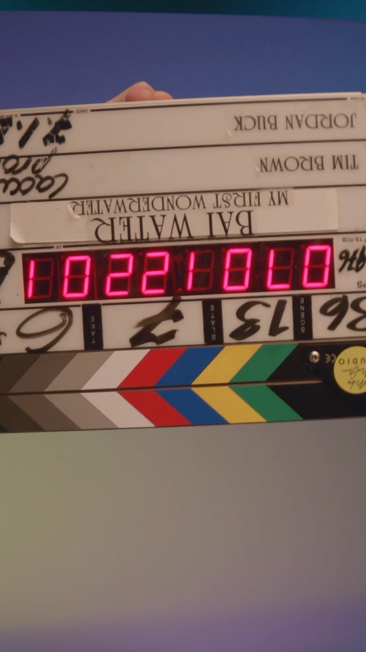 a closeup shot of a clapperboard held by a hand.  the clapperboard displays the following information:  "8513" and "002200" as scene and take numbers,  "bai water" as the title of the production, "my first wonderwater" as a subtitle, "tim brown" and "jordan buck" as names. the color bars are visible at the bottom. the background is a blurry light blue.