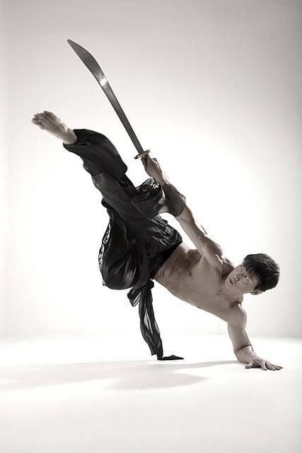 a man in black clothing is performing a martial arts move with a sword. he is in midair, legs extended, and holding the sword with one hand. he is looking at the camera with an intense expression. the background is white.