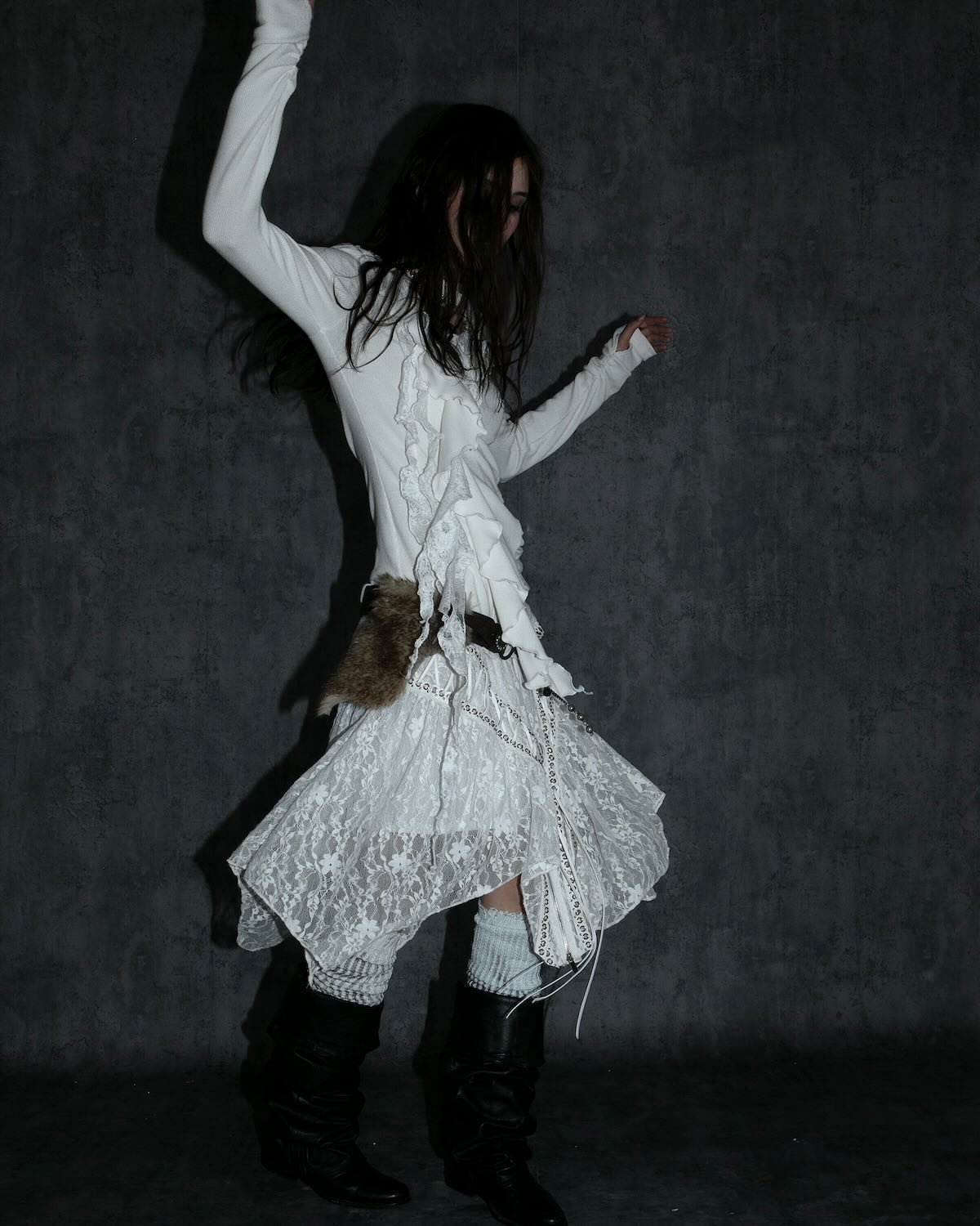 a full shot of a woman modeling a white lace dress with ruffles and a layered skirt.  she's wearing high black boots and has long dark hair. the background is a dark, textured grey. the overall style is dark, artistic, and slightly gothic. the image has a moody and stylish feel.