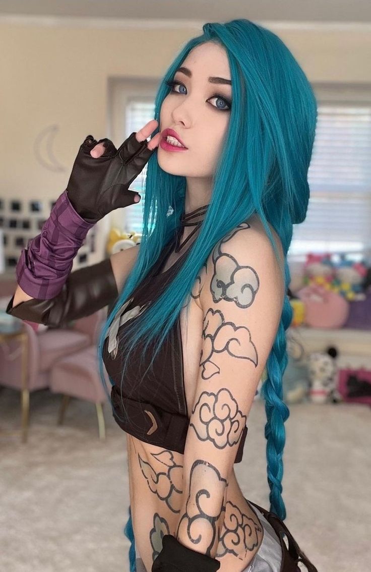 a woman cosplaying as jinx from league of legends stands indoors. she has long blue hair styled in a braid, and is wearing fingerless gloves and a brown crop top. she has tattoos of cloudlike designs on her arms. she is holding her fingers near her lips in a playful gesture.