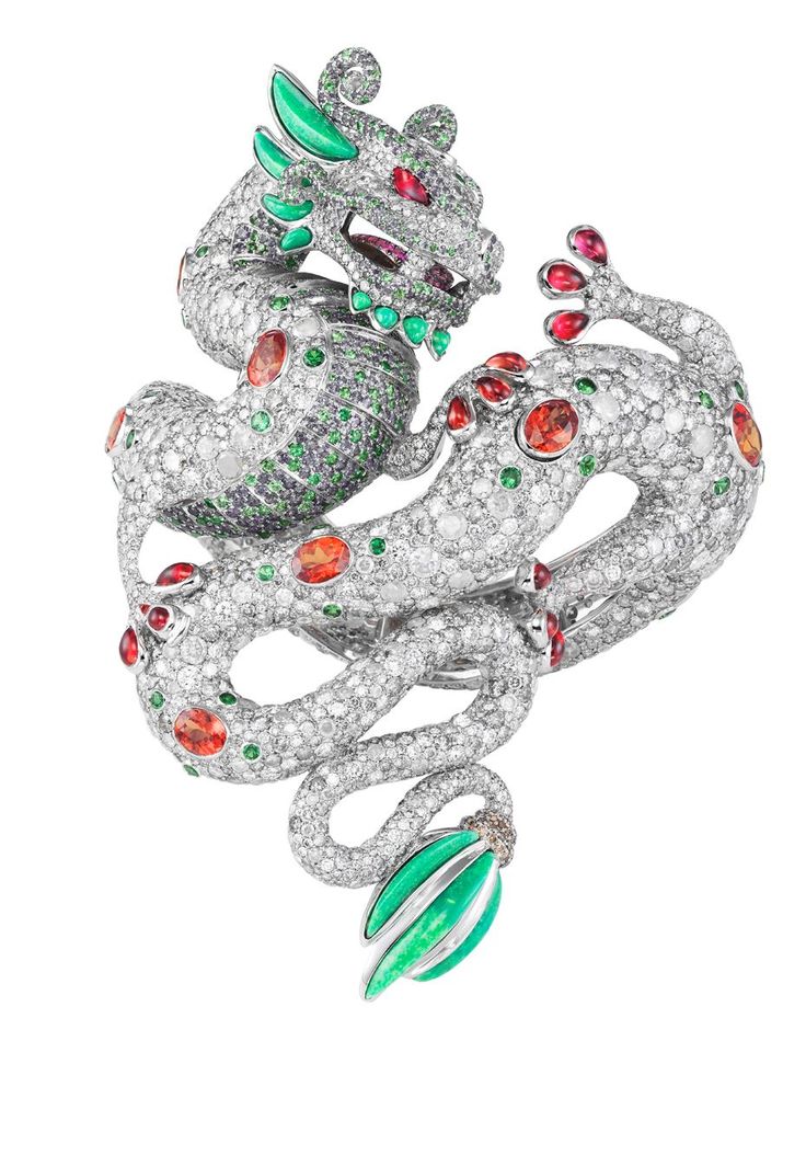 an ornate dragon brooch made of white gold and encrusted with diamonds and gemstones. the dragon has red eyes, green horns and accents, and a body covered in small diamonds. it is posed in a coiled position and features a green and white gold tail.
