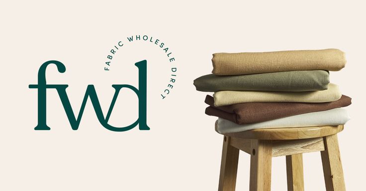 the image shows a logo for fabric wholesale direct (fwd) next to a stack of folded fabrics on a wooden stool. the fwd logo is in dark green and next to it, in a curved formation, are the words "fabric wholesale direct". on the right, a small wooden stool holds a stack of beige, green, brown, and white fabrics. the background is a plain light beige color.