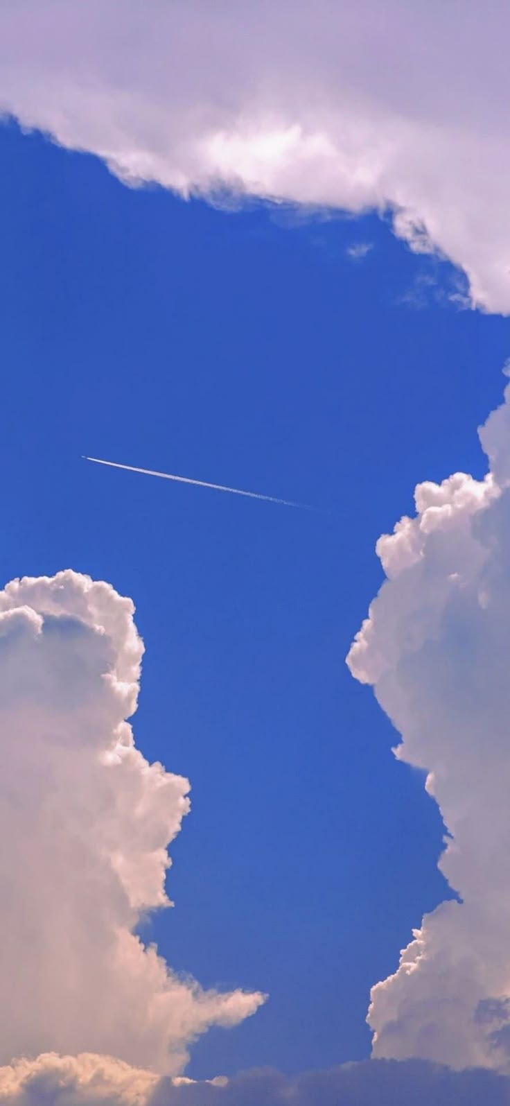 a vertical shot of a bright blue sky framed by large, fluffy white clouds on either side, with a long white contrail stretching across the center of the open sky.