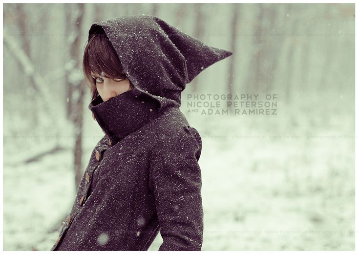 a young woman with dark hair and a dark gray coat with a pointed hood stands in a snowy forest. the photo is taken from an eyelevel perspective, with the woman facing to the left of the frame and slightly towards the camera. the background is blurred, creating a sense of depth and emphasizing the falling snow. the overall palette is muted and cool, with shades of gray, white, and brown.