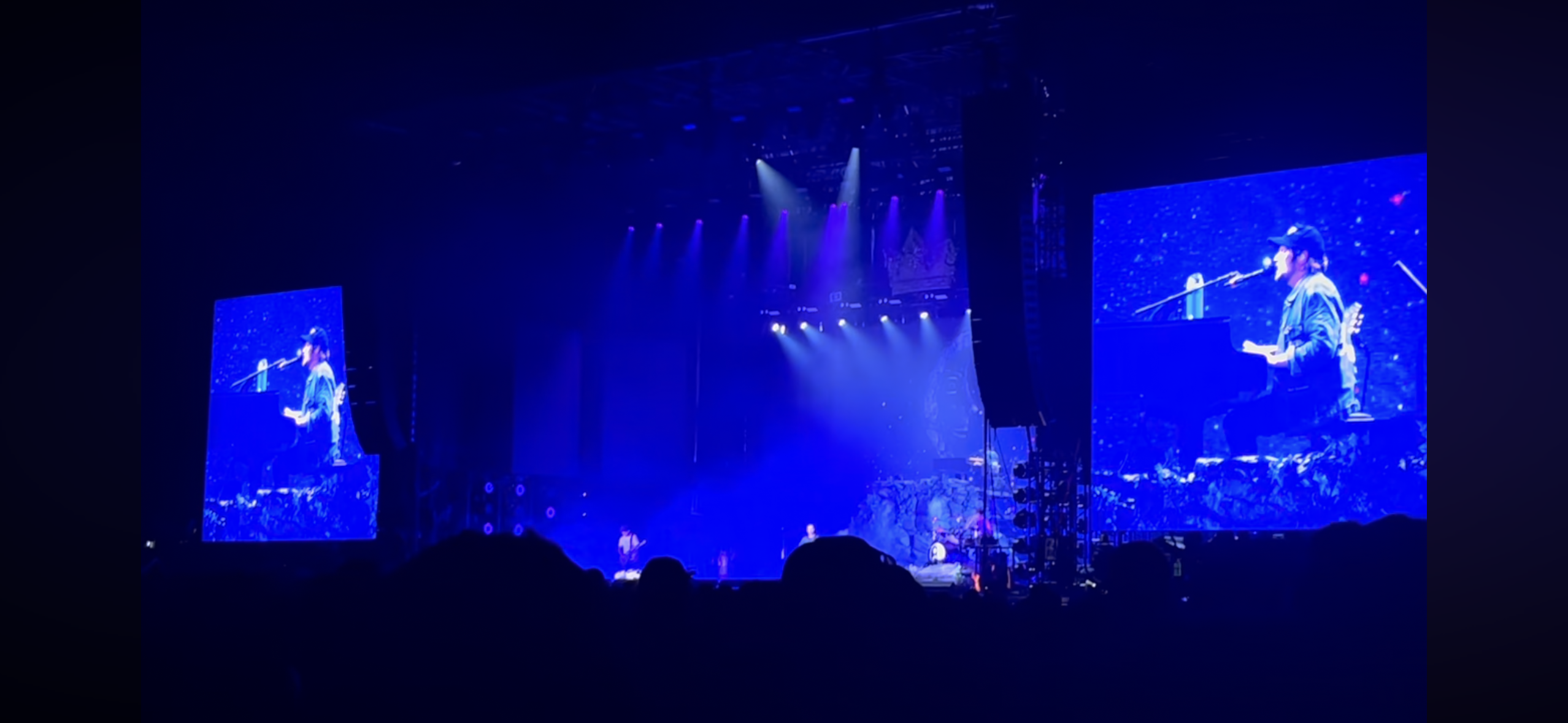 a concert stage is lit with blue lights. the stage is dark and there are two large screens that show a musician playing piano and singing. there are other silhouettes of band members in the background. the crowd is blacked out in the foreground. the image is taken from a concert venue.