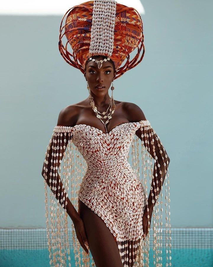 a stunning darkskinned woman is captured in a portrait wearing a unique and elaborate outfit. she has a large, intricately designed headdress adorned with a pattern and vertical white band of cowrie shells. she wears a matching offtheshoulder dress also covered in cowrie shells. a long waterfall of cowrie shells drapes from her sleeves. she is also wearing multiple necklaces and dangling earrings. the woman is standing in front of a light blue wall and a pool of water, creating a bright and visually appealing backdrop. the focus is on the woman and her striking appearance.