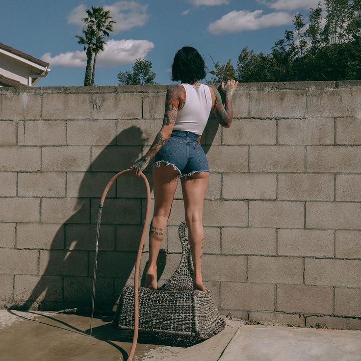a woman with tattoos, wearing jean shorts and a white tank top, stands with her back to the camera. she is holding a hose that is spraying water onto the ground. she is standing on a large woven basket and leaning against a cinderblock wall. the scene appears to be a backyard on a sunny day.