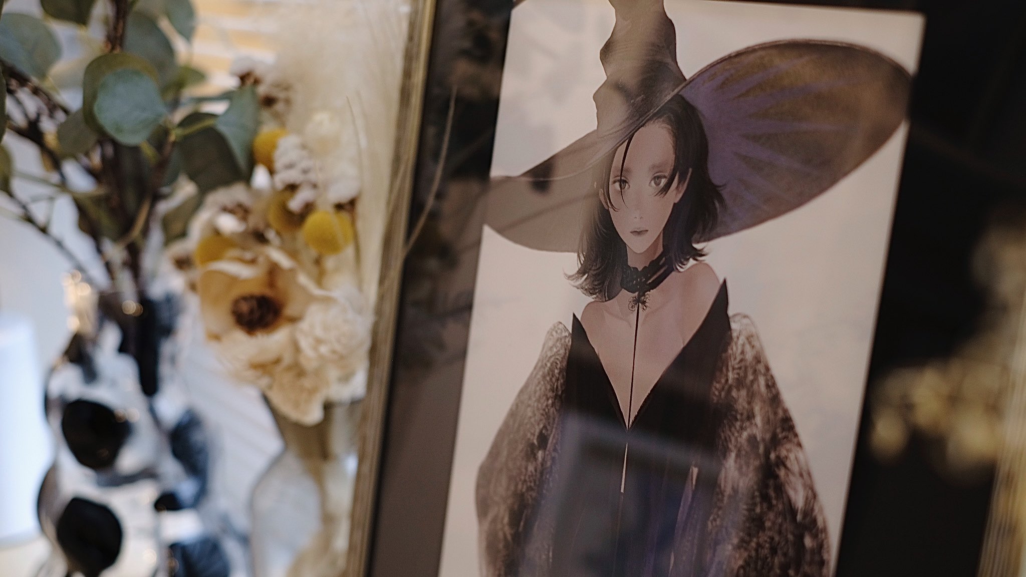 the image shows a framed print of a woman dressed as a witch. the woman in the portrait has pale skin, dark hair, and is wearing a large black hat and a black and grey velvet dress.  the frame is black and is sitting on a surface next to a vase of flowers and greenery. the lighting in the image is soft and diffused.