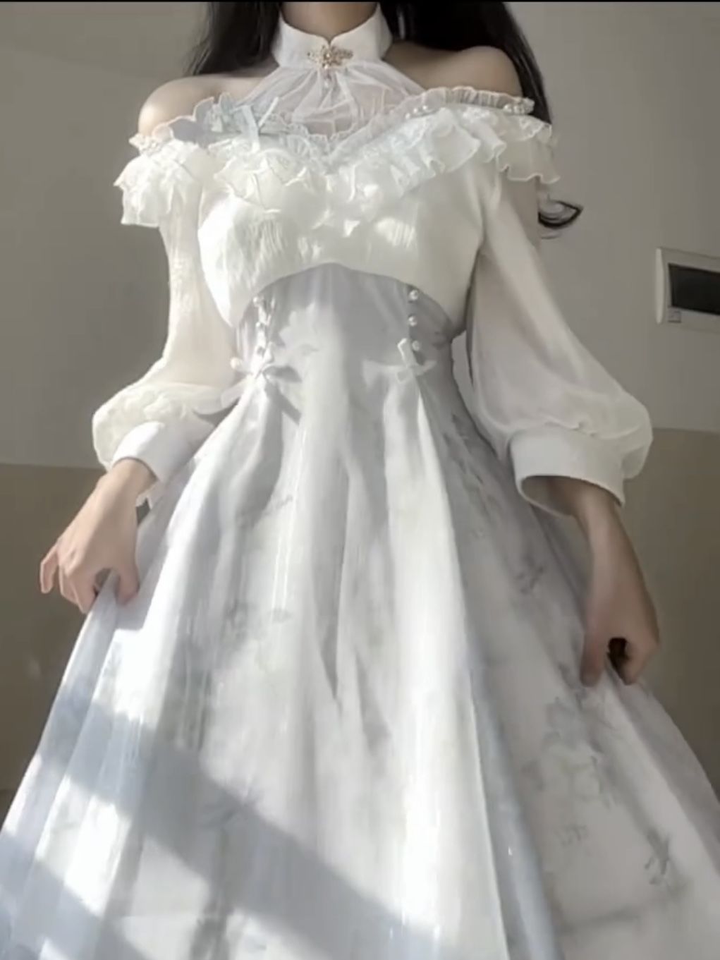 a person wearing a white and blue offshoulder gown with long sleeves and a high collar is standing in front of a wall. the dress has ruffles around the neckline and a pattern on the skirt. the fabric is sheer and looks vintage. the person is holding the dress with both hands. a brooch is pinned to the high collar.