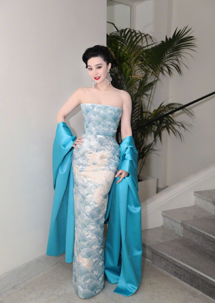 a woman in a beautiful blue and teal gown with a strapless design and a long train is standing on a staircase. she is smiling and looking at the camera. she has black hair and a natural, elegant look. she is accessorized with large earrings. the background is white and the stair steps are made of marble. she is giving a gentle pose and looking at the camera with a smile on her face.  the dress is heavily embellished with lace and sequins, and the fabric has a shimmer. this is a formal event, such as a gala or a party.