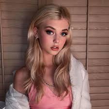 the image features a young woman with blonde hair and blue eyes, wearing a pink top and a white jacket. she is posing indoors against a backdrop of blinds. she has fair skin and is wearing makeup. the image is a portrait.