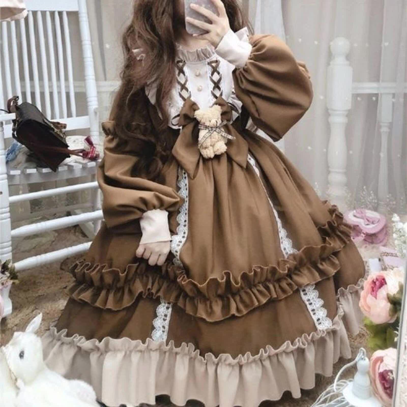 a young woman is wearing a brown and cream lolita fashion dress. the dress has a square neckline, long sleeves, and a full skirt with ruffles and lace trim. the bodice is decorated with a bow and a teddy bear charm. the woman is standing in a room with white walls and a wooden chair. there are some flowers and a white stuffed animal on the floor.