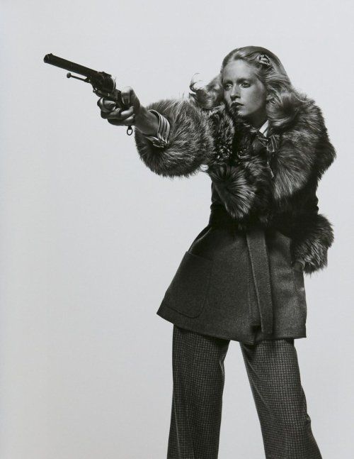 a woman in a fur coat and pants poses in a studio, holding a pistol with her right hand. she has long blond hair and a serious expression on her face. the photo is in black and white, giving it a vintage look.