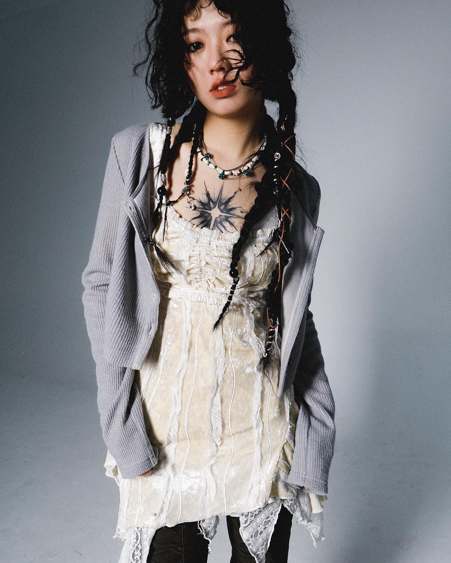 a young woman with dark hair styled in braids poses for a studio portrait. she's wearing a grey ribbed cardigan over an offwhite velvet dress with lace detailing.  she has several necklaces and a noticeable chest tattoo. the overall style is edgy and alternative. the background is a plain light grey.