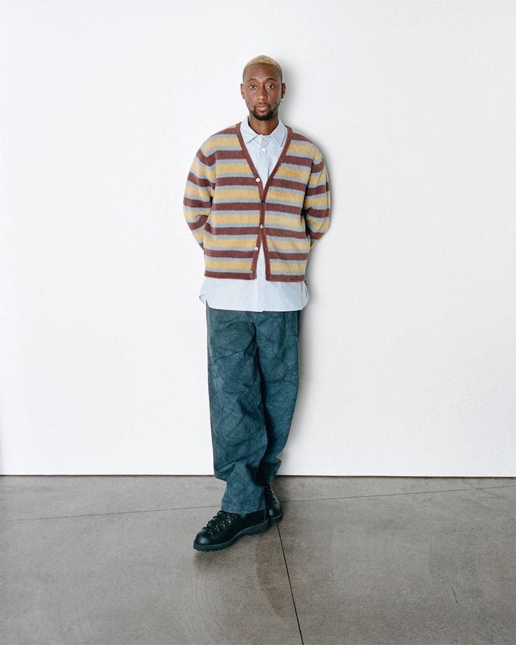 a fullbody shot of a man standing against a white wall. he's wearing a striped brown and beige cardigan over a light blue buttondown shirt and dark blue jeans. his black boots are visible. the setting is minimalistic and clean. the man's pose is relaxed and casual. the overall image presents a clean, modern fashion look.
