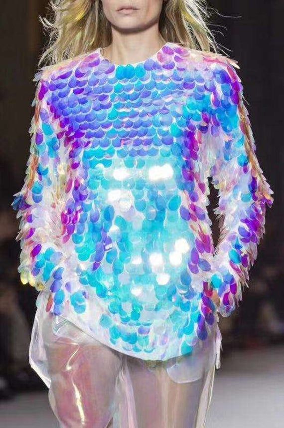 a woman is modeling an iridescent sequined dress that shimmer in a variety of colors, including white, blue, pink, and purple. the dress is formfitting and features long sleeves. she is standing on a runway, and her expression is one of confidence. the dress is both elegant and stylish, making her look stunning. the sequins catch the light beautifully, giving the dress a mesmerizing effect.