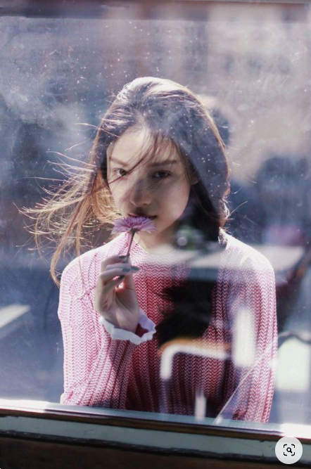 a young woman with long dark hair wearing a pink sweater is seen through a window. she is holding a small pink flower up to her face and smelling it. her reflection can be seen in the window. the lighting is soft and natural, and the overall tone of the image is dreamy and romantic.