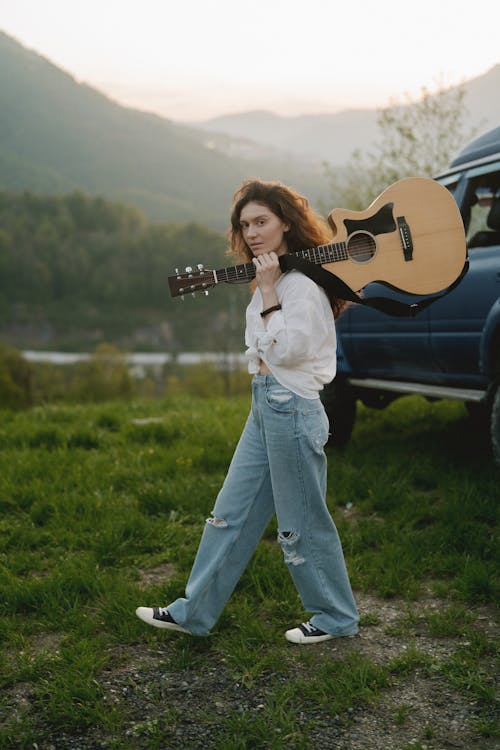a young woman with curly brown hair walks across a grassy field, carrying an acoustic guitar on her shoulder.  she's wearing a white blouse and loosefitting, ripped jeans.  a darkcolored van is parked in the background, partially visible, against a backdrop of rolling green hills and a clear sky. the overall atmosphere is relaxed and peaceful, suggesting a journey or adventure. the image is brightly lit, possibly during the golden hour of sunset or sunrise.