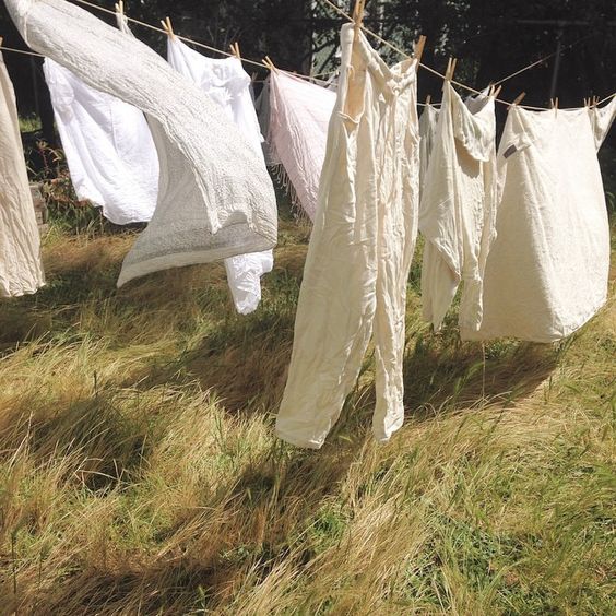a line of white linen clothes is drying on a clothesline in a field of tall grass on a sunny day. the wind is blowing gently, and the clothes are billowing in the breeze. the scene is peaceful and serene, with a rustic, vintage feel.