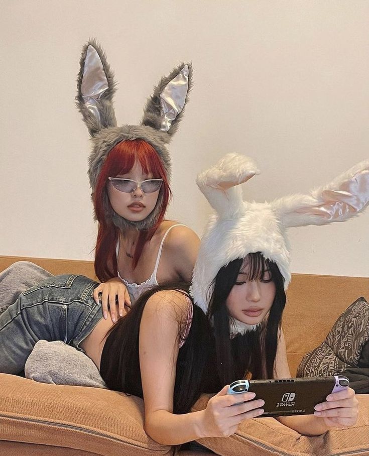 two girls are laying on a couch, one wearing a white rabbit ear hat and the other wearing a grey bunny ear hat. both girls are looking at the camera and smiling. the girl in the white bunny ear hat is holding a nintendo switch and playing a video game.